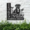 Black metal house sign on white brick showing number 1565 and text FAIRWAY DRIVE with a sitting dog, flowers and butterflies, greenery below