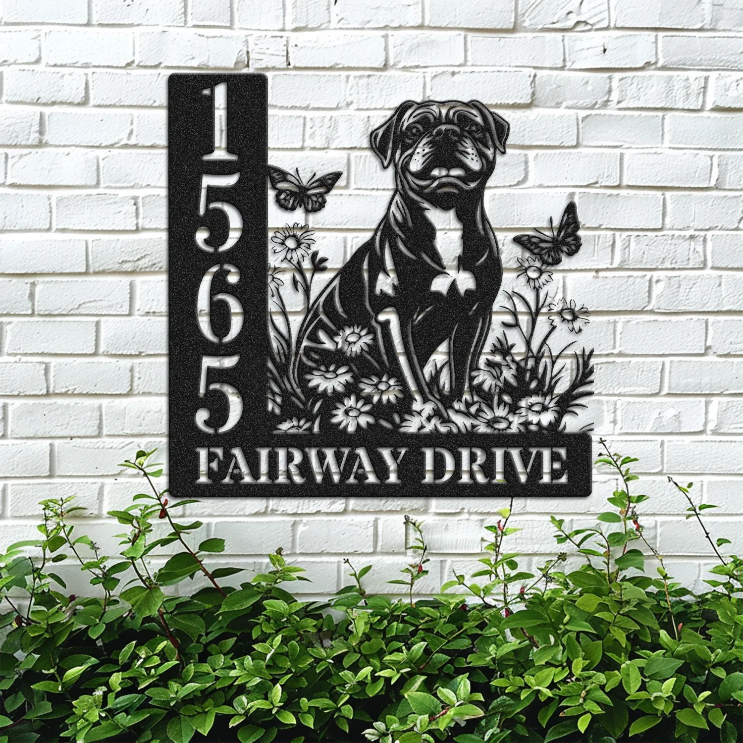 Black metal house sign on white brick showing number 1565 and text FAIRWAY DRIVE with a sitting dog, flowers and butterflies, greenery below