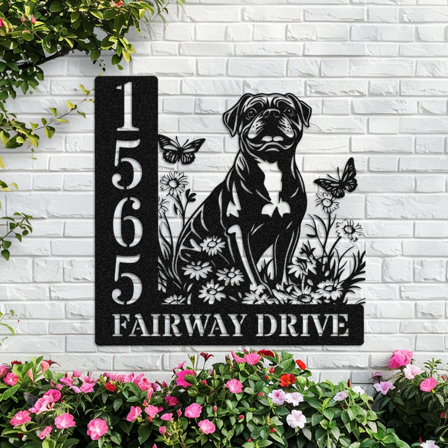Black metal house sign with address numbers 1565 and text FAIRWAY DRIVE, featuring a sitting dog silhouette, flowers and butterflies mounted on a white brick wall above pink flowering bushes