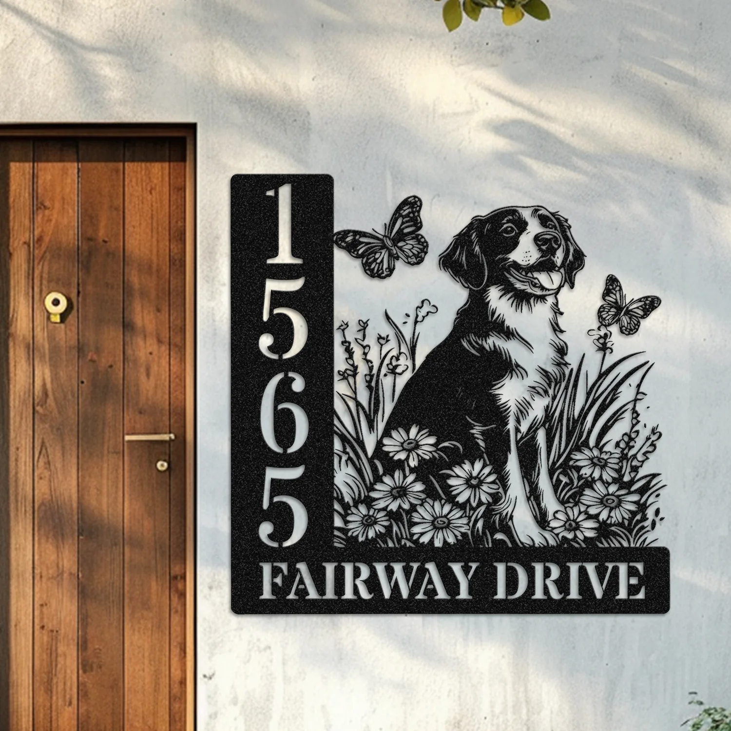 Black metal house sign mounted on a light exterior wall showing address numbers 1565, text 'FAIRWAY DRIVE', and a dog with flowers and butterflies next to a wooden door.