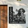 Black metal house sign mounted on an exterior wall next to a wooden door showing numbers 1565 and the text FAIRWAY DRIVE with a dog, flowers and butterflies design