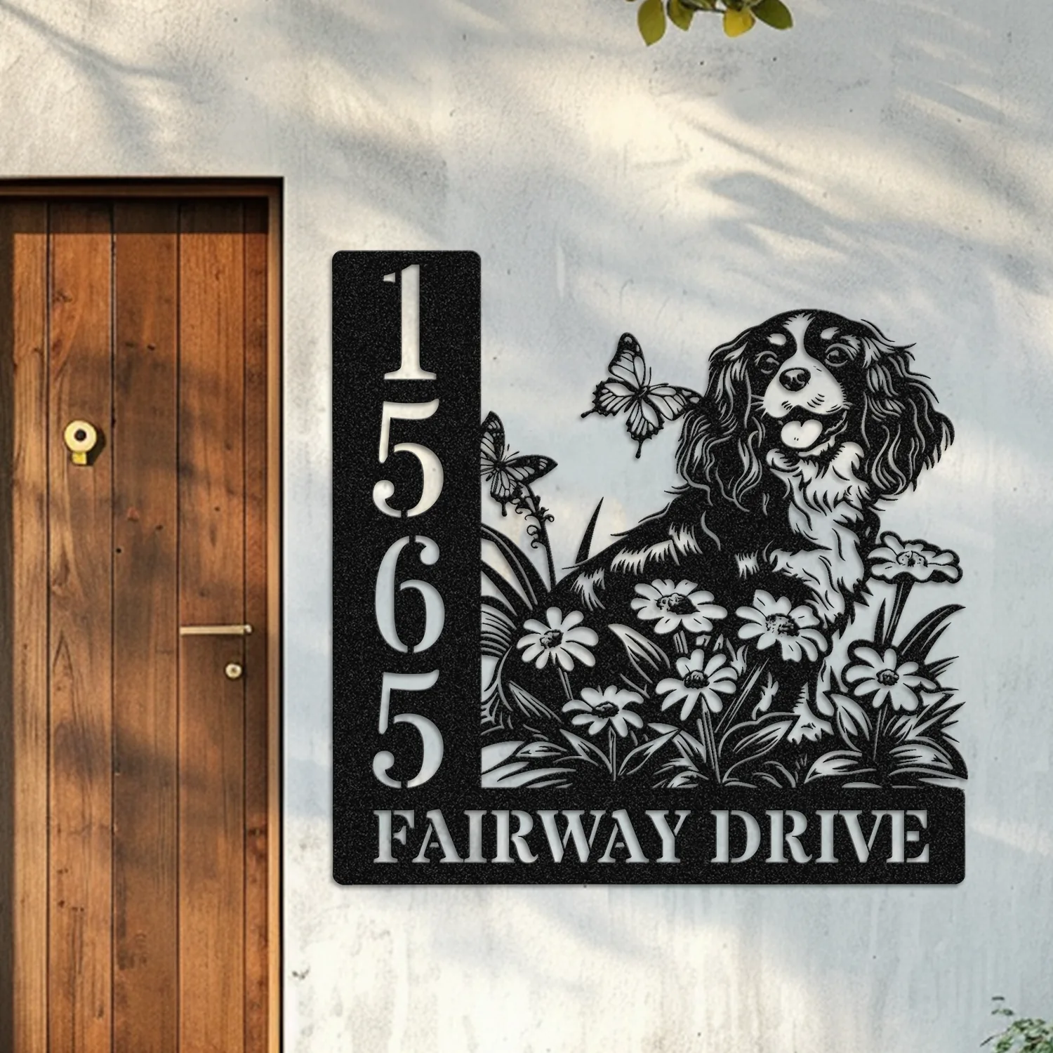 Cavalier-King-Charles-Spaniel-Sitting-with-Butterfly-1-2 Black metal house sign mounted beside a wooden door showing address numbers 1565 and text “FAIRWAY DRIVE” with a dog, flowers and butterflies design