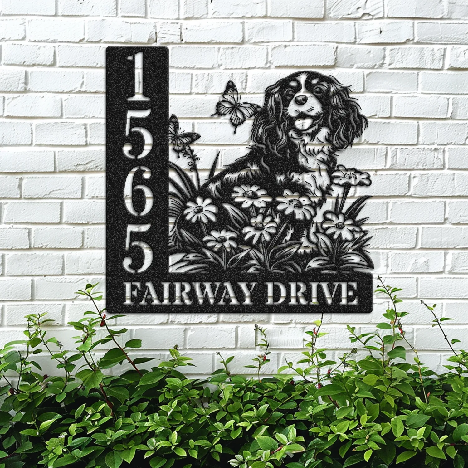 Black metal house sign mounted on a white brick wall showing numbers 1565 and text "FAIRWAY DRIVE" with a dog, flowers and butterflies, greenery below