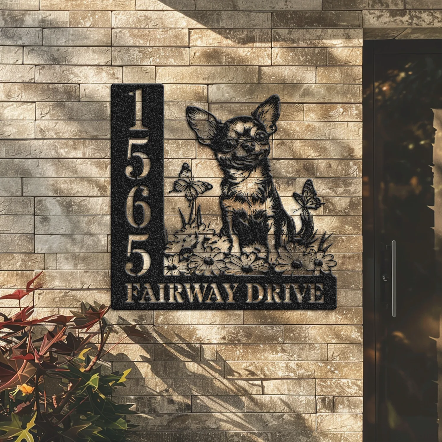 Black metal house sign mounted on a tan brick wall showing numbers 1565 and text 'FAIRWAY DRIVE' with a Chihuahua silhouette among flowers and butterflies