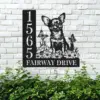 Black metal house sign mounted on a white brick wall showing number 1565 and the words FAIRWAY DRIVE with a Chihuahua sitting among flowers and butterflies above green shrubs