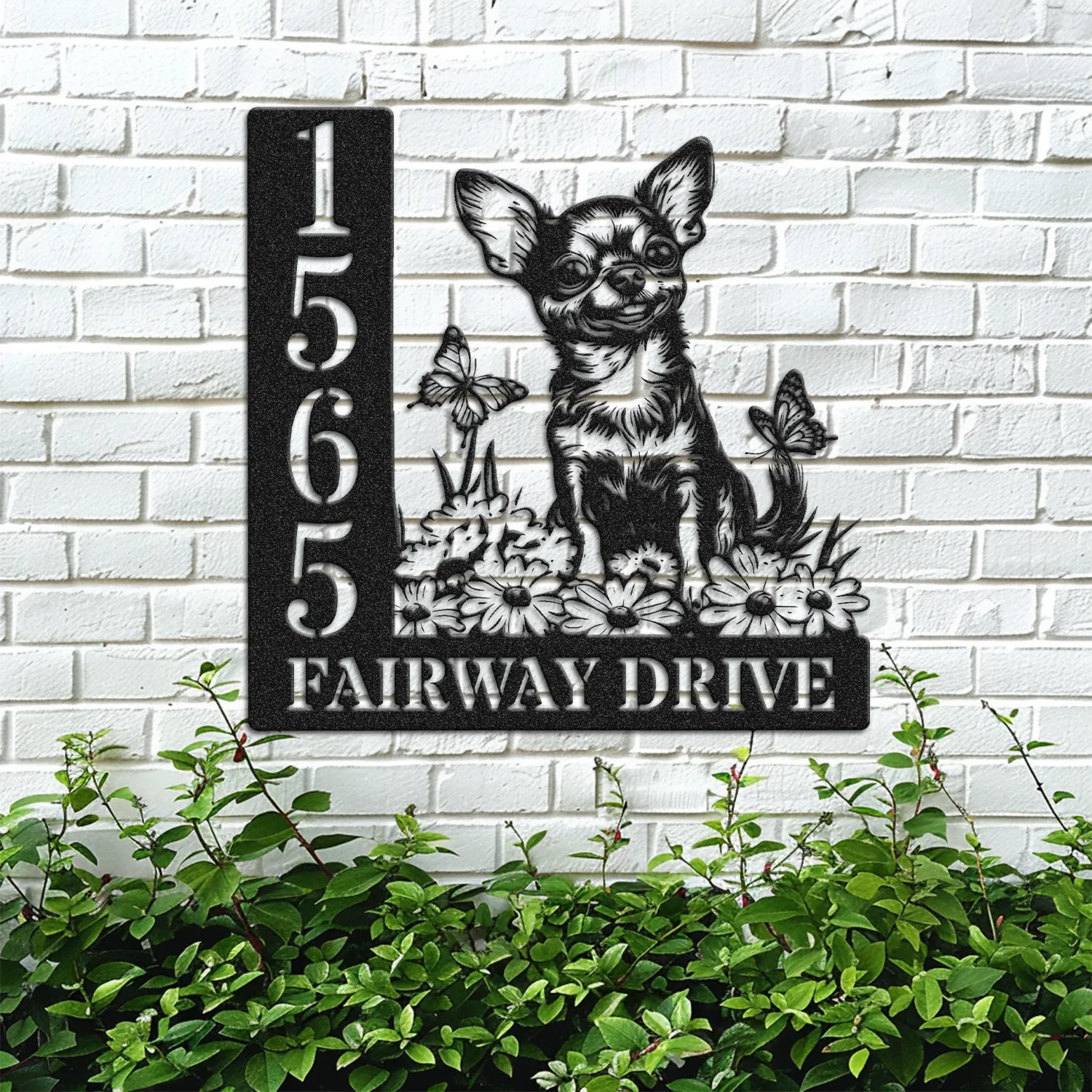 Black metal house sign mounted on a white brick wall showing number 1565 and the words FAIRWAY DRIVE with a Chihuahua sitting among flowers and butterflies above green shrubs
