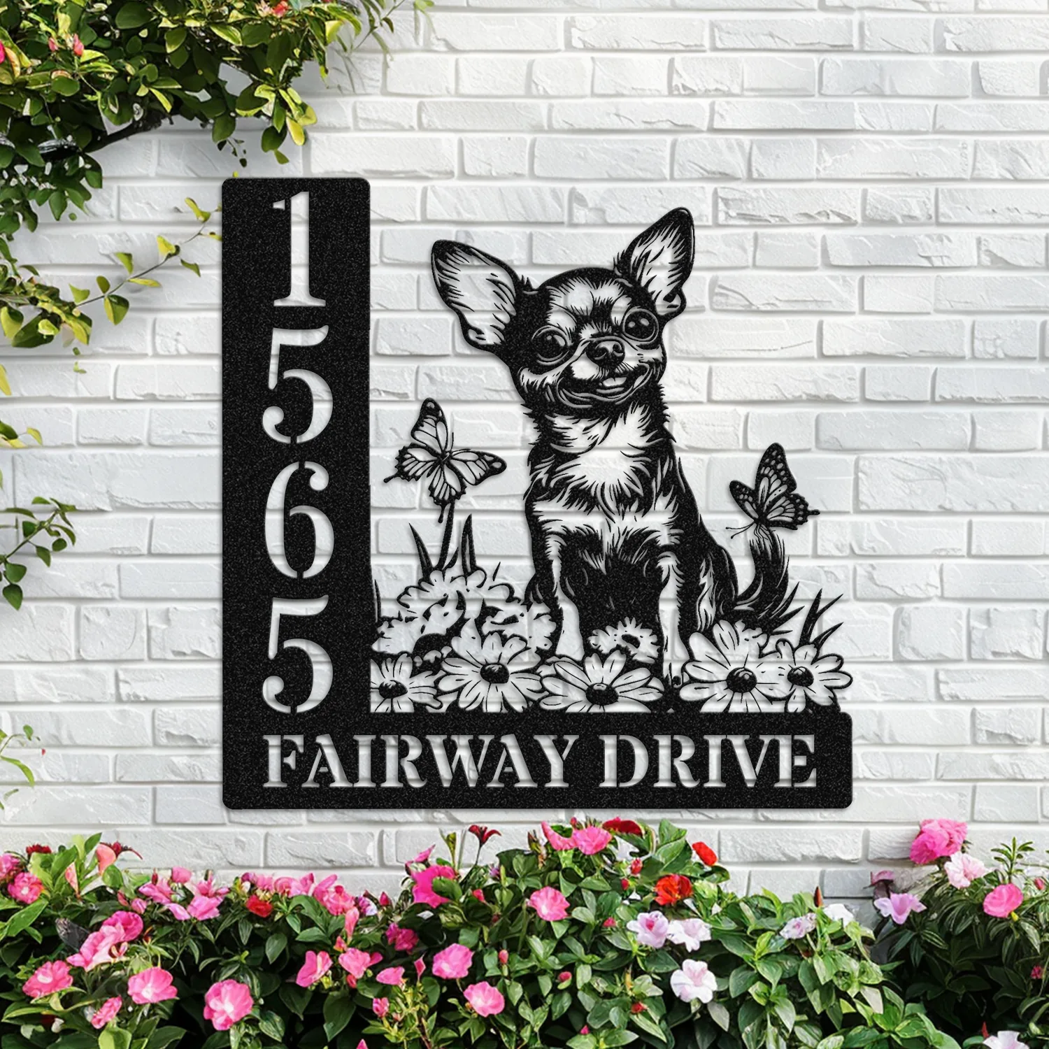 Black metal house number sign mounted on a white brick wall with numbers 1 5 6 5 and text 'FAIRWAY DRIVE' beside a decorative cutout of a chihuahua, flowers and butterflies, with pink flowers below