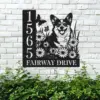 Black metal house sign mounted on white brick showing numbers 1565, text 'FAIRWAY DRIVE', daisies, butterflies and a smiling corgi among greenery
