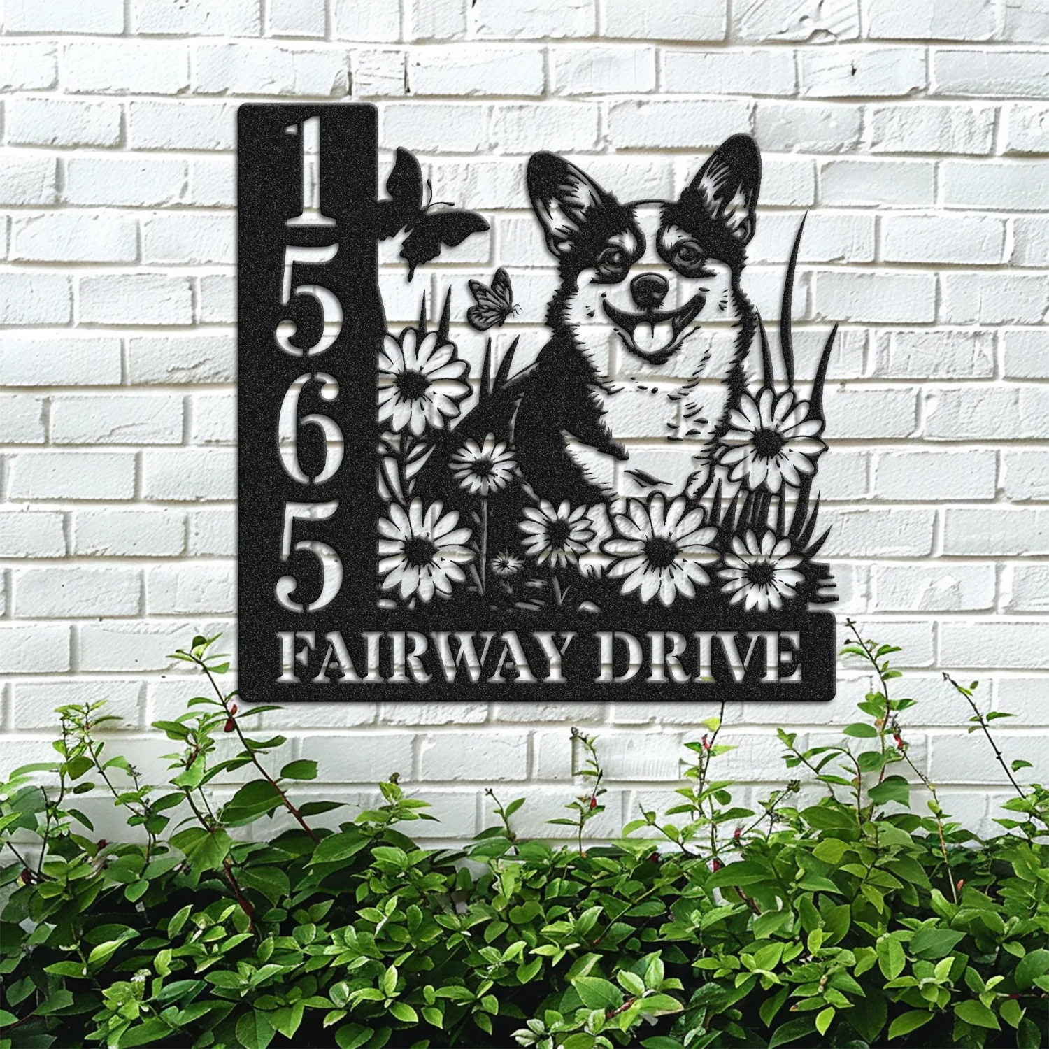 Black metal house sign mounted on white brick showing numbers 1565, text 'FAIRWAY DRIVE', daisies, butterflies and a smiling corgi among greenery