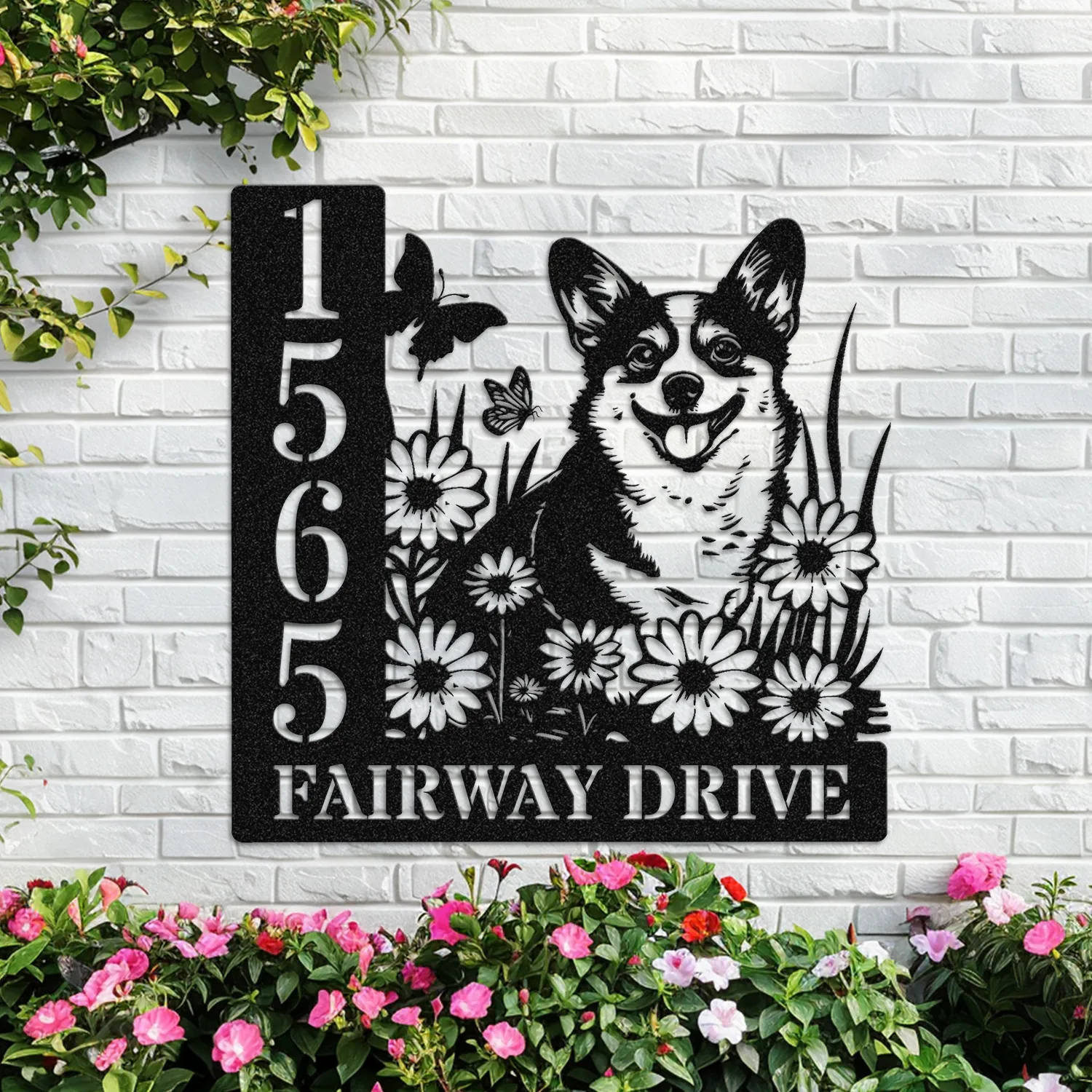Corgi-Sitting-with-Butterfly-4 Black metal address sign mounted on a white brick wall showing numbers 1565, text ‘FAIRWAY DRIVE’, a smiling corgi among daisies with butterflies, surrounded by flowering plants