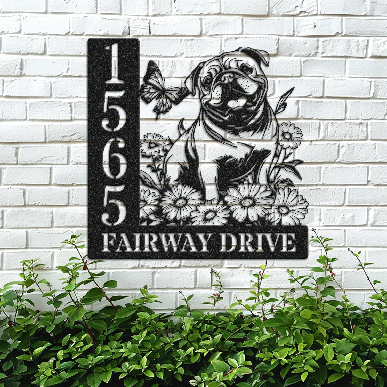 English-Bulldog-Sitting-with-Butterfly-3 Black metal house number sign reading 1565 and FAIRWAY DRIVE with a cutout pug, flowers and butterfly mounted on a white brick wall above green shrubbery
