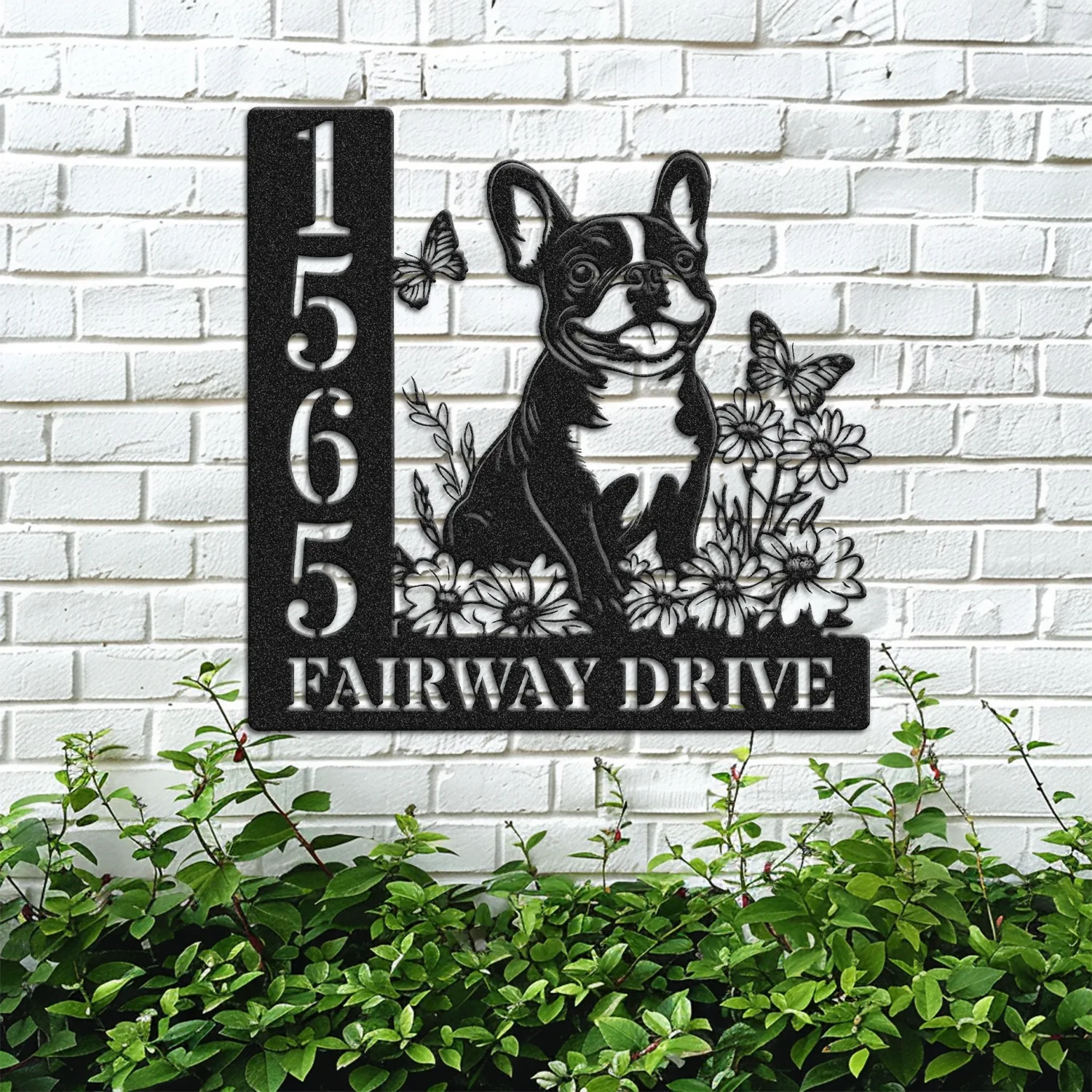 French-Bulldog-Sitting-with-Butterfly-2 Black metal house sign mounted on a white brick wall showing address 1565 and text FAIRWAY DRIVE with a sitting French bulldog, flowers, and butterflies above green shrubs.