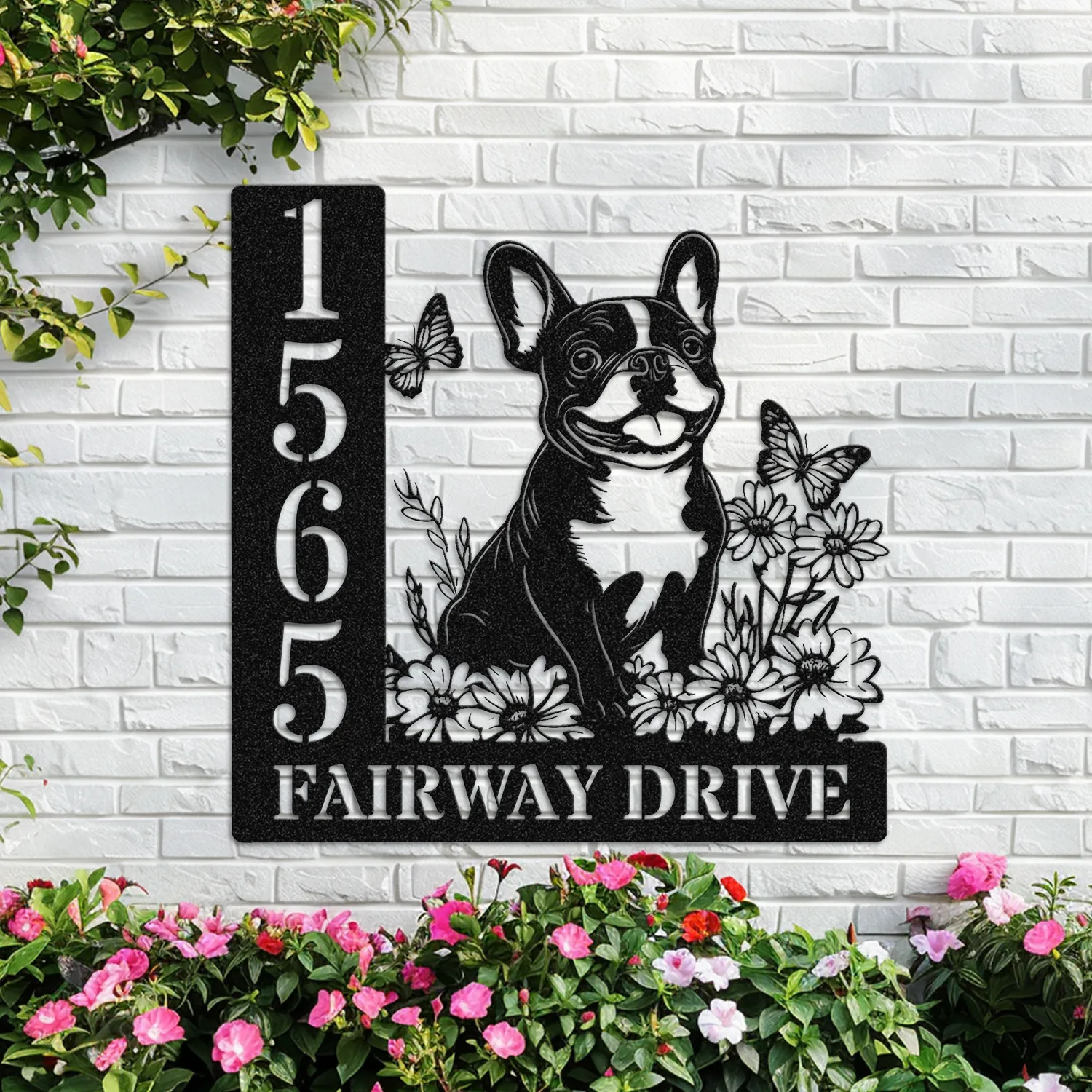 French-Bulldog-Sitting-with-Butterfly-3 Black metal house sign mounted on a white brick wall showing number 1565, a smiling French bulldog amid flowers and butterflies, text 'FAIRWAY DRIVE' and pink flowers below