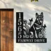 Black metal house sign mounted on a white exterior wall next to a wooden door showing numbers 15655, text 'FAIRWAY DRIVE', and a German shepherd with flowers and butterflies