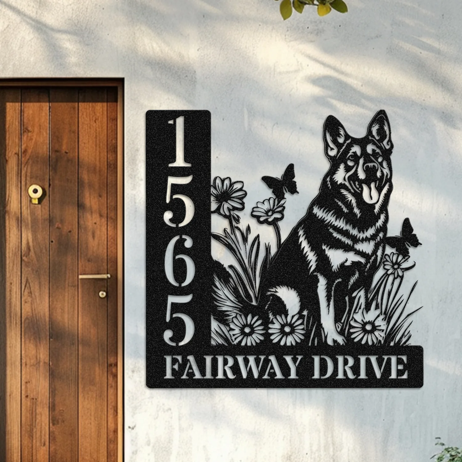Black metal house sign mounted on a white exterior wall next to a wooden door showing numbers 15655, text 'FAIRWAY DRIVE', and a German shepherd with flowers and butterflies