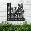 Black metal house sign mounted on a white brick wall showing numbers 1565 and the words FAIRWAY DRIVE with a German shepherd silhouette among flowers and butterflies above green shrubs
