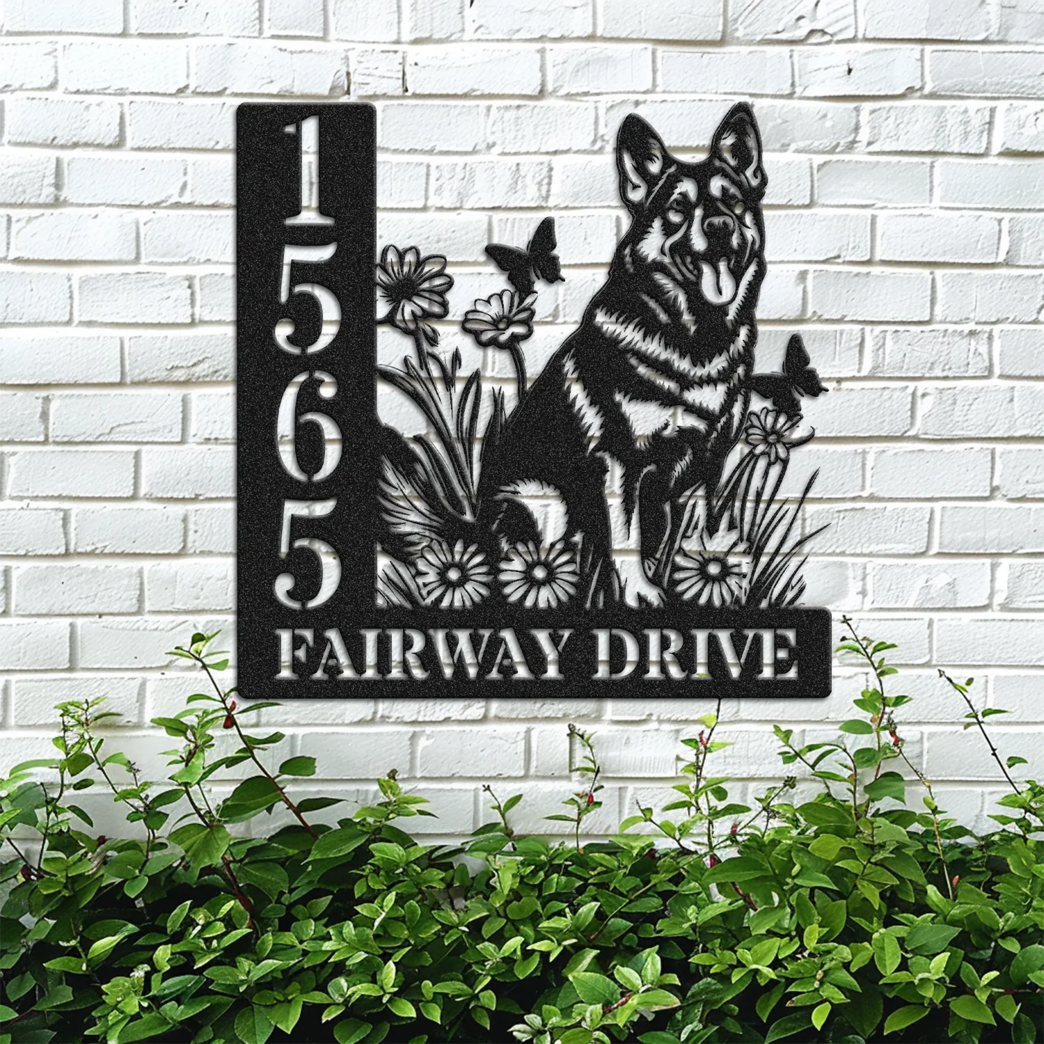 Black metal house sign mounted on a white brick wall showing numbers 1565 and the words FAIRWAY DRIVE with a German shepherd silhouette among flowers and butterflies above green shrubs