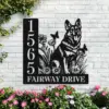 Black metal house sign on a white brick wall showing numbers 1 5 6 5 and text 'FAIRWAY DRIVE' with a German Shepherd silhouette, flowers and butterflies, planted flowers below