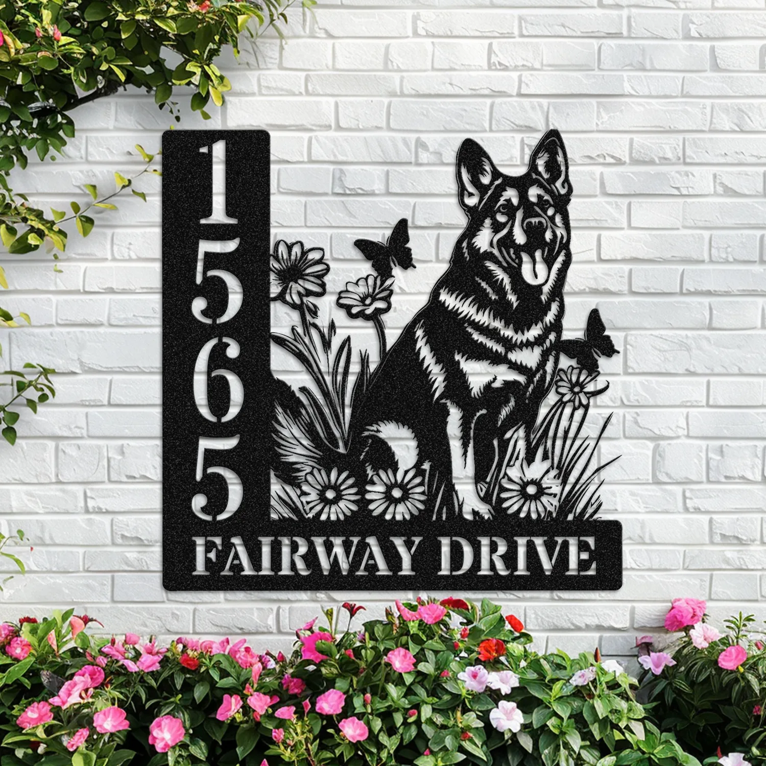 Black metal house sign on a white brick wall showing numbers 1 5 6 5 and text 'FAIRWAY DRIVE' with a German Shepherd silhouette, flowers and butterflies, planted flowers below