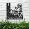 Black metal house sign mounted on a white brick wall showing numbers 1565 and text FAIRWAY DRIVE beside a sitting dog and flowers, greenery below