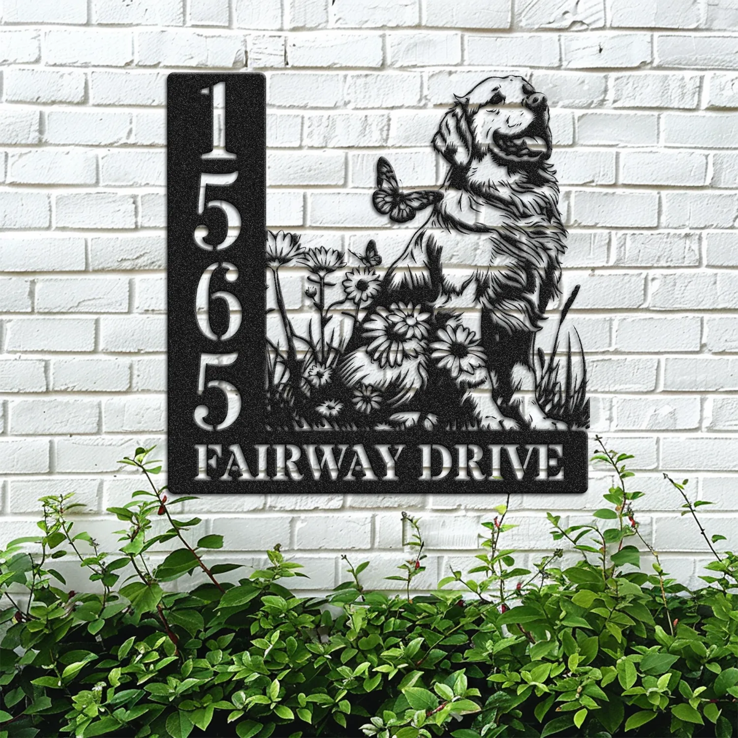 Black metal house sign mounted on a white brick wall showing numbers 1565 and text FAIRWAY DRIVE beside a sitting dog and flowers, greenery below