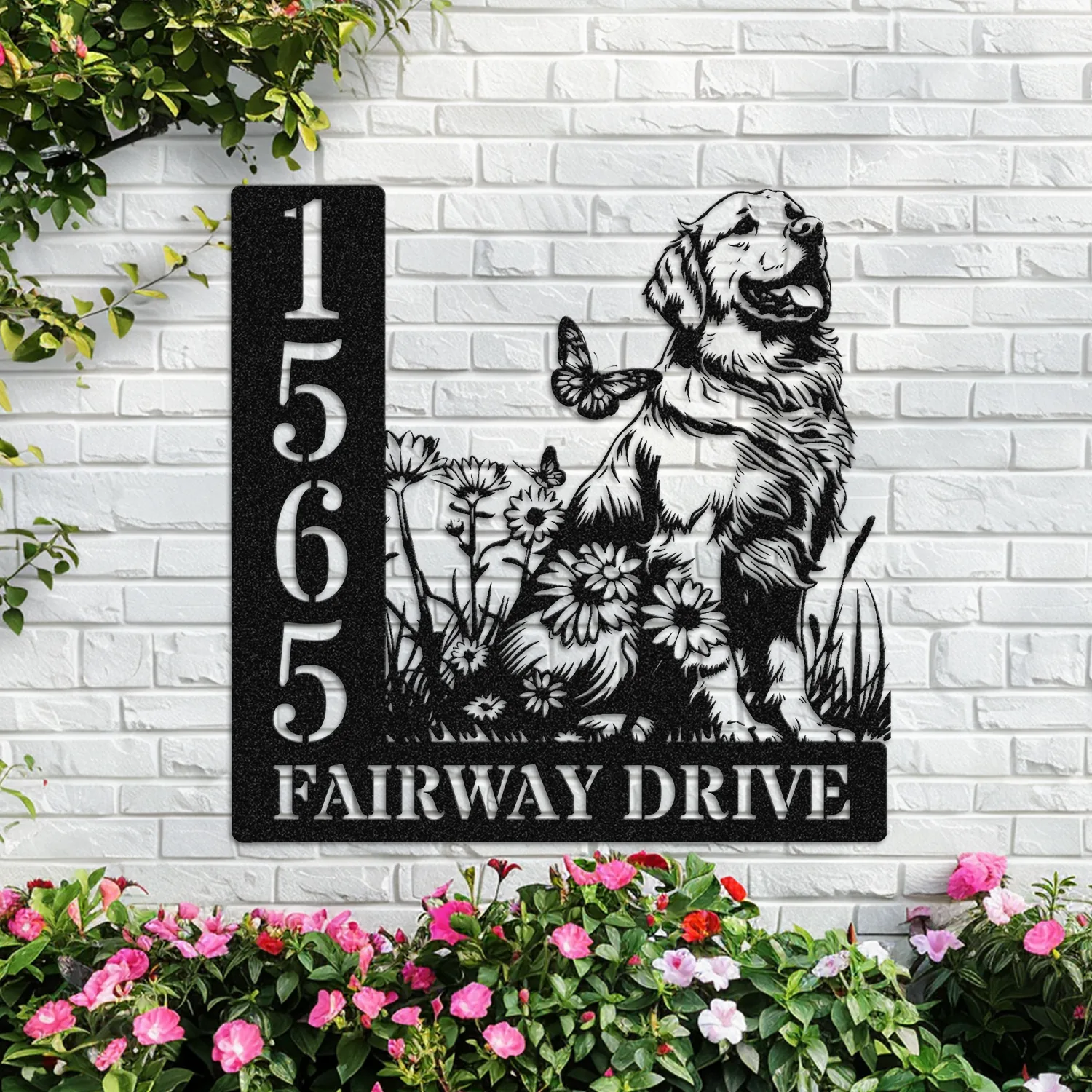 black metal house sign mounted on a white brick wall showing address 1565 and the text FAIRWAY DRIVE with a sitting golden retriever and flowers, pink garden blooms below