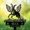 Black memorial sign with winged dog silhouette leaping above flowers, text "In Loving Memory Of" and name MAX with dates 2016-2024 on a sunlit grassy background