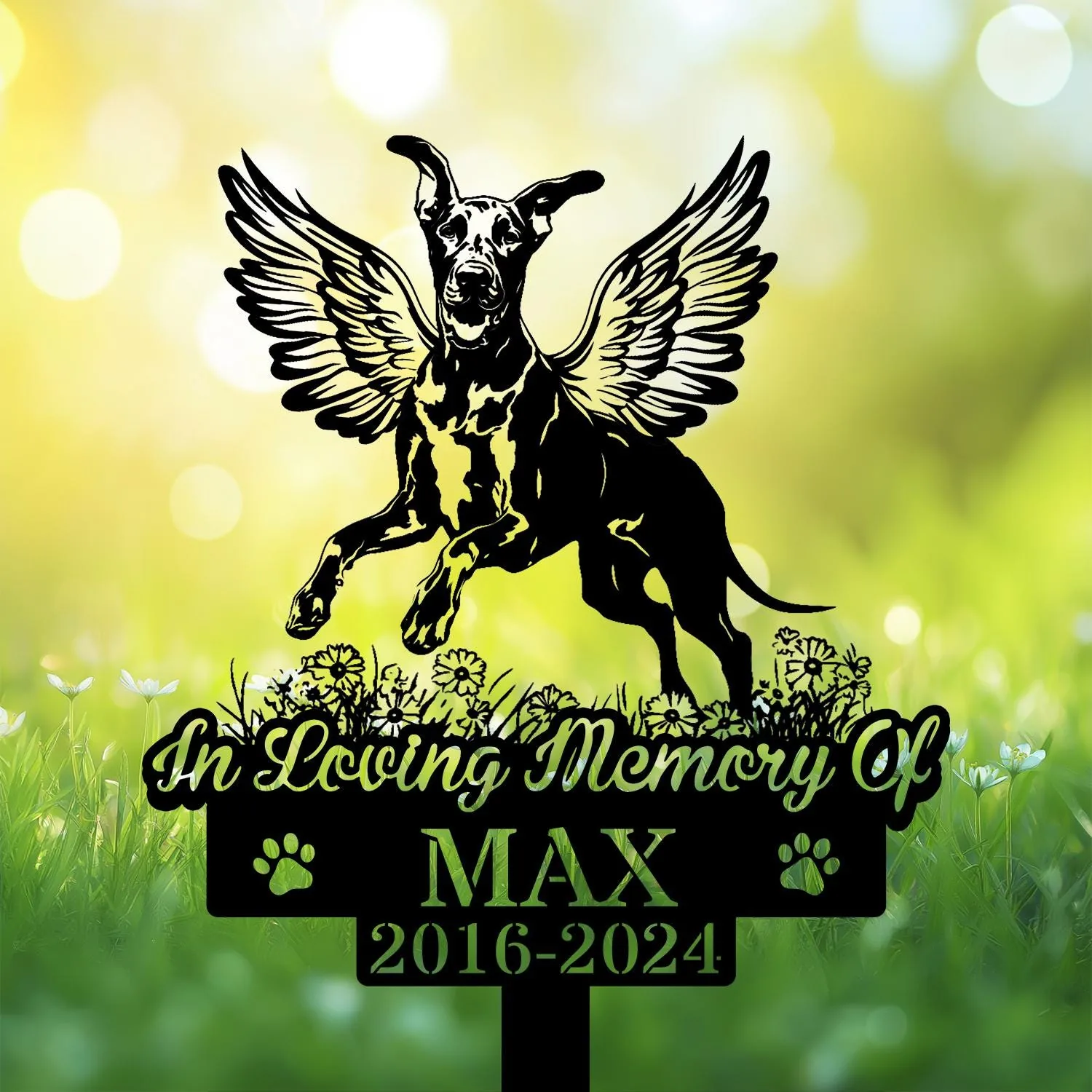Black memorial sign with winged dog silhouette leaping above flowers, text "In Loving Memory Of" and name MAX with dates 2016-2024 on a sunlit grassy background