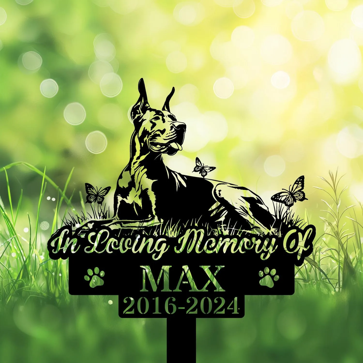 Memorial garden sign showing a reclining dog silhouette with butterflies, text "In Loving Memory Of MAX 2016-2024" against a sunlit green bokeh background