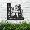 Black metal house sign on a white brick wall showing numbers 1565 and text FAIRWAY DRIVE with a sitting golden retriever, flowers and butterflies above green shrubs