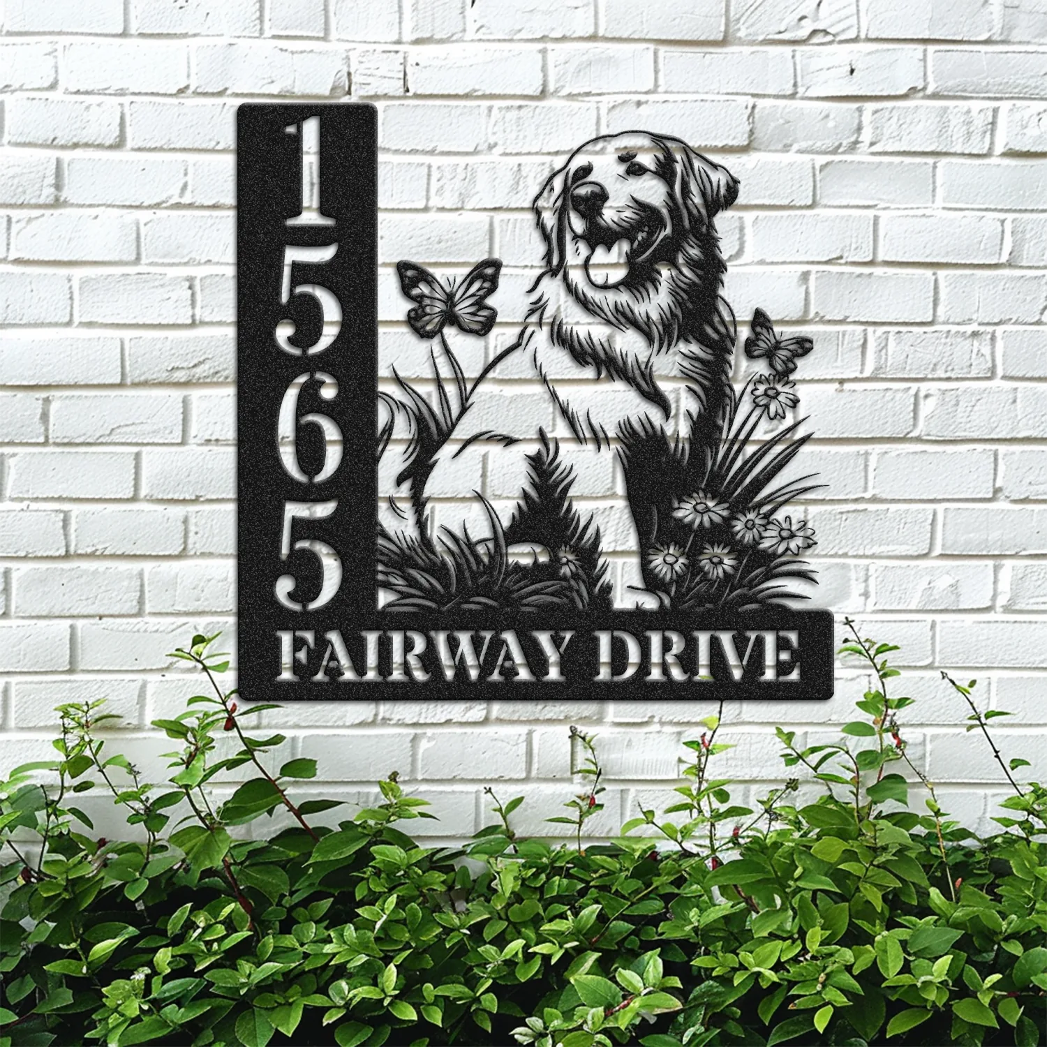 Black metal house sign on a white brick wall showing numbers 1565 and text FAIRWAY DRIVE with a sitting golden retriever, flowers and butterflies above green shrubs