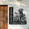 Black metal house sign mounted next to a wooden door showing vertical address 1565 and text ‘FAIRWAY DRIVE’ with floral artwork, butterflies and a sitting dog