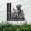 Black metal house sign mounted on a white brick wall showing numbers 1565 and the text FAIRWAY DRIVE with a dog, flowers and butterflies design above green bushes