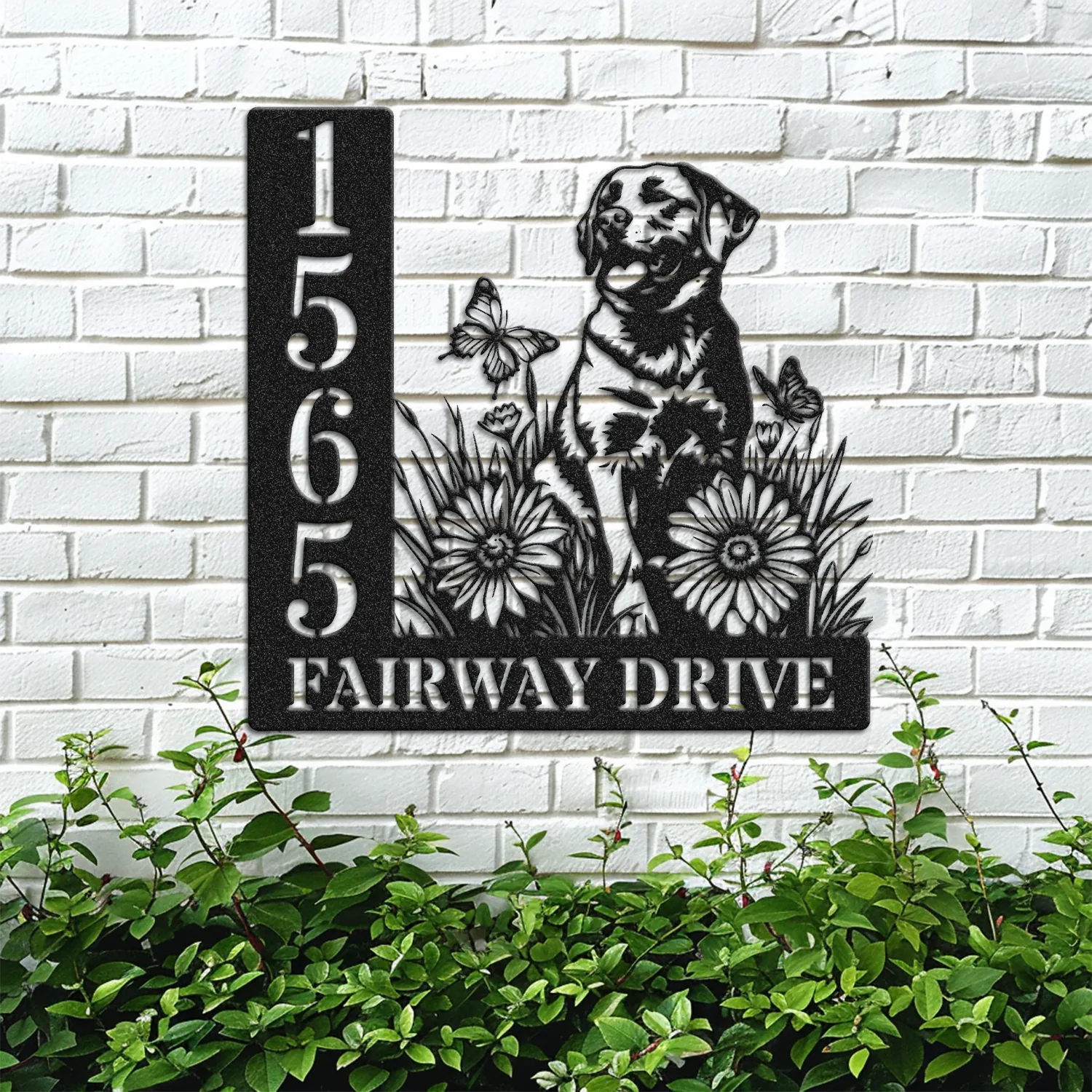 Black metal house sign mounted on a white brick wall showing numbers 1565 and the text FAIRWAY DRIVE with a dog, flowers and butterflies design above green bushes