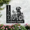 Black metal address sign mounted on white brick wall showing numbers 1565 and text 'FAIRWAY DRIVE' with a dog, flowers and butterflies, pink flowers in front