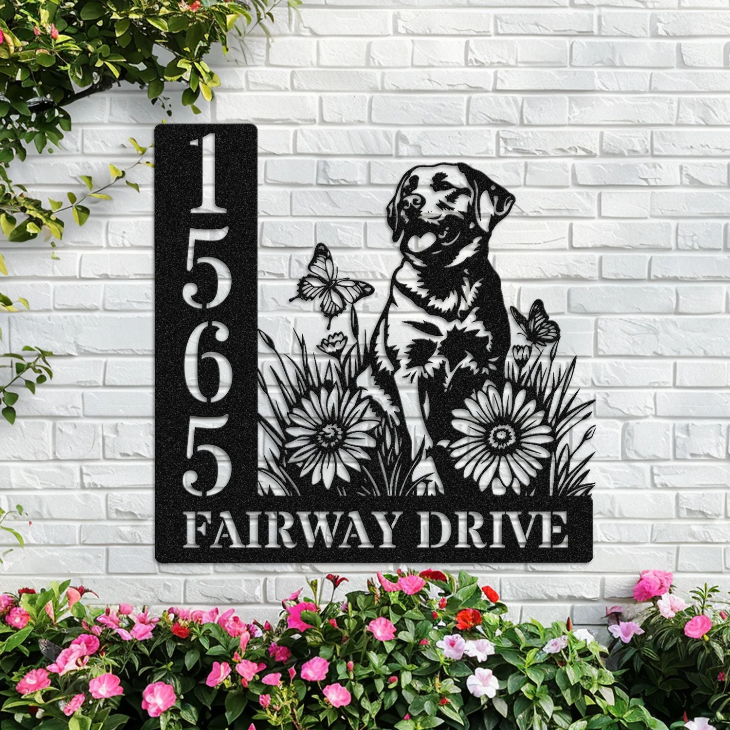 Black metal address sign mounted on white brick wall showing numbers 1565 and text 'FAIRWAY DRIVE' with a dog, flowers and butterflies, pink flowers in front