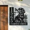 Black metal address sign mounted on a light stucco wall beside a wooden door, showing numbers 15655 and text 'FAIRWAY DRIVE' with a dog, flowers and butterfly design