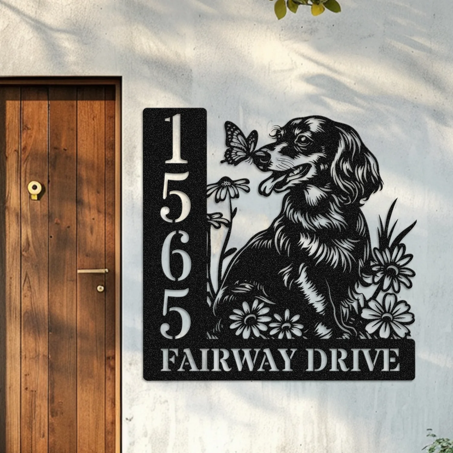 Black metal address sign mounted on a light stucco wall beside a wooden door, showing numbers 15655 and text 'FAIRWAY DRIVE' with a dog, flowers and butterfly design