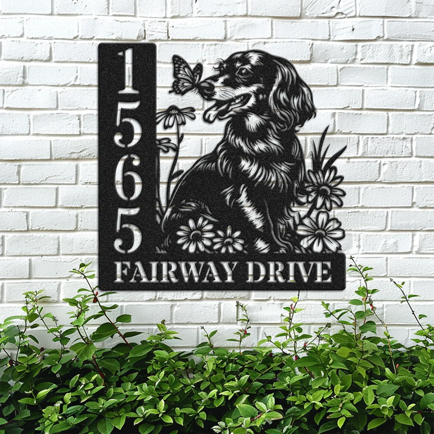 Black metal house sign mounted on a white brick wall showing number 1565 and text "Fairway Drive" with a dog, flowers, and a butterfly design above green shrubbery