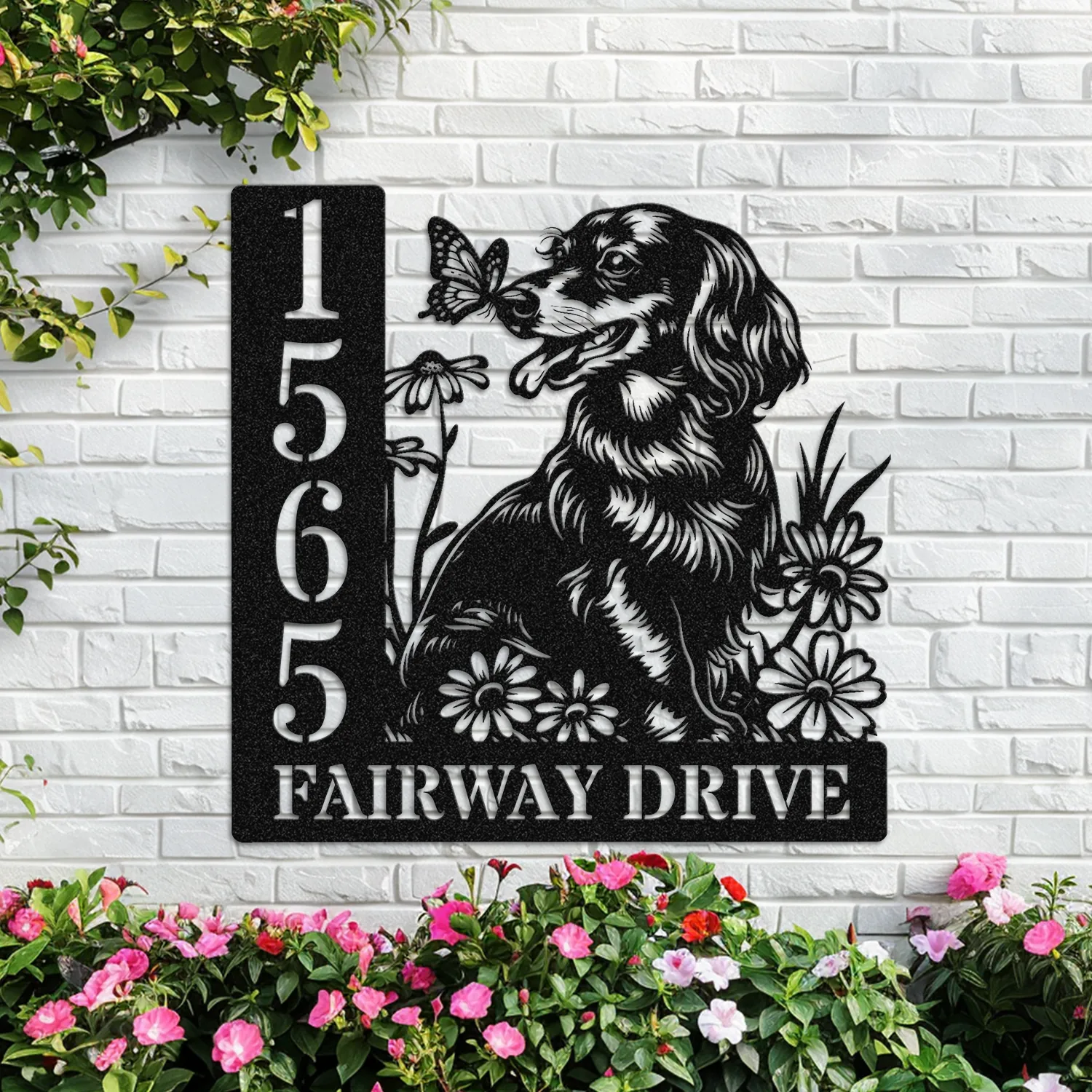 Black metal house sign mounted on a white brick wall featuring a dog with a butterfly, daisies, vertical house number 1565 and text 'FAIRWAY DRIVE' with pink flowers at the base