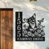 Black metal address sign mounted on a white exterior wall next to a wooden door, showing numbers 1565 and text 'FAIRWAY DRIVE' with a happy dog surrounded by daisies and butterflies