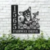 Black metal house sign mounted on a white brick wall showing address 1565 and text FAIRWAY DRIVE with a cheerful dog, flowers and butterflies above green shrubbery