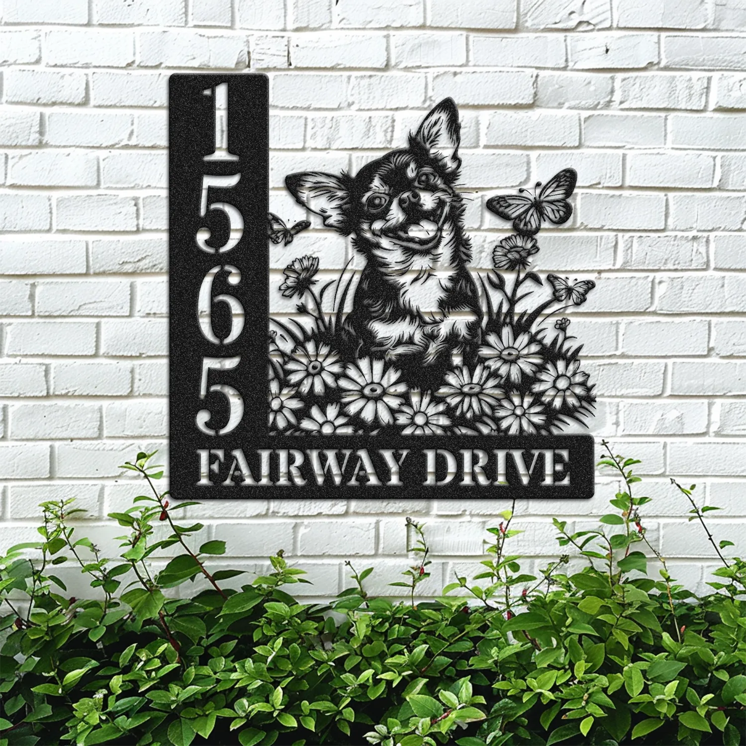 Black metal house sign mounted on a white brick wall showing address 1565 and text FAIRWAY DRIVE with a cheerful dog, flowers and butterflies above green shrubbery