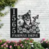 Black metal house sign mounted on a white brick wall showing address numbers and text 'FAIRWAY DRIVE' with a happy dog illustration, daisies and butterflies, flowering plants below