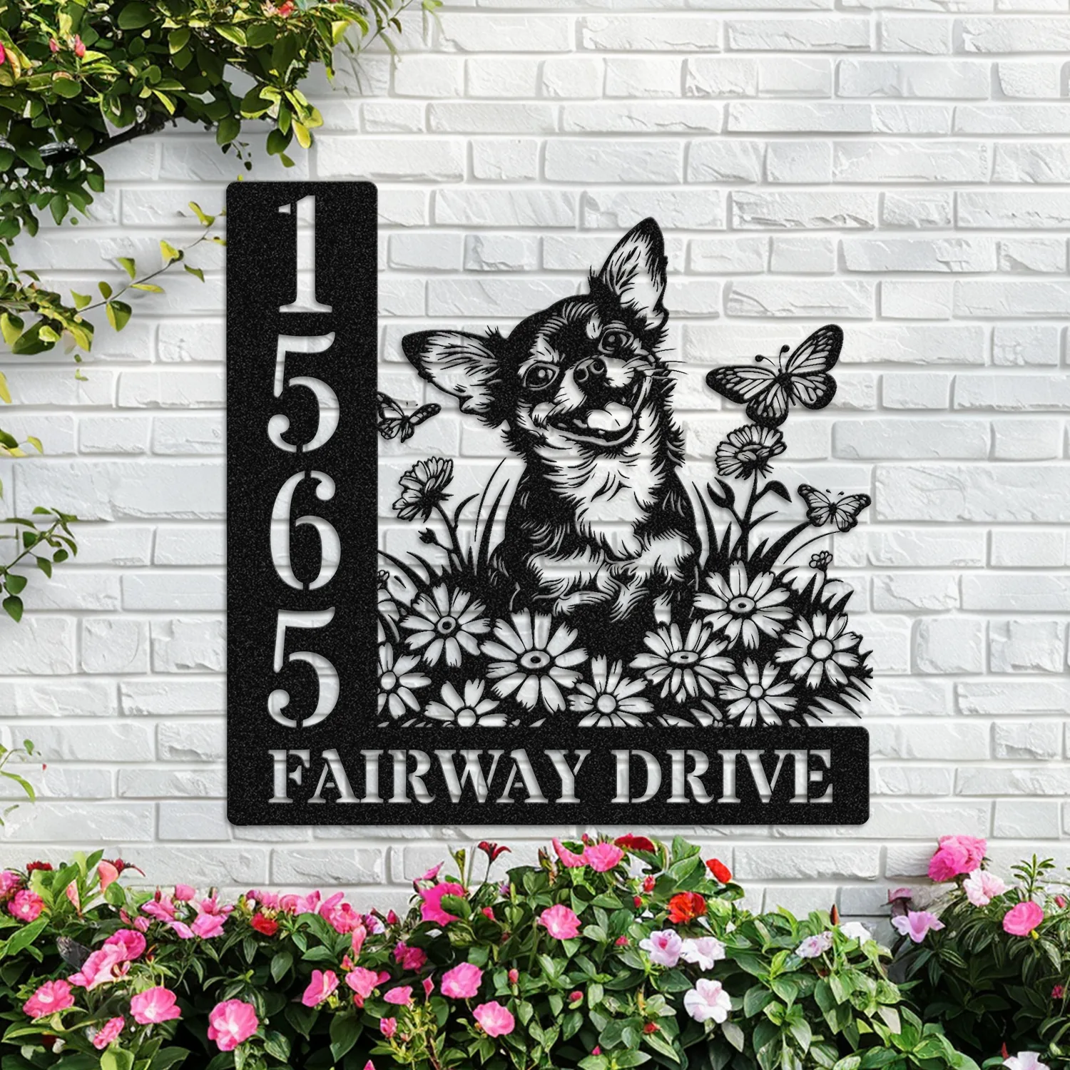 Black metal house sign mounted on a white brick wall showing address numbers and text 'FAIRWAY DRIVE' with a happy dog illustration, daisies and butterflies, flowering plants below