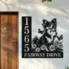 Black metal house sign mounted next to a wooden door with vertical address numbers 1565 and text FAIRWAY DRIVE beside a cheerful long-haired dog among flowers and butterflies