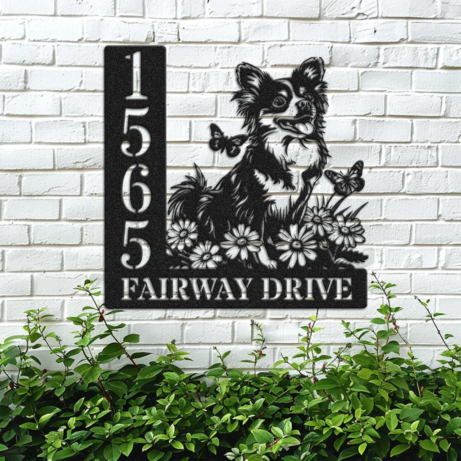 Black metal house sign mounted on a white brick wall showing numbers 1565 and text FAIRWAY DRIVE with a cheerful dog, flowers and butterflies above green shrubs
