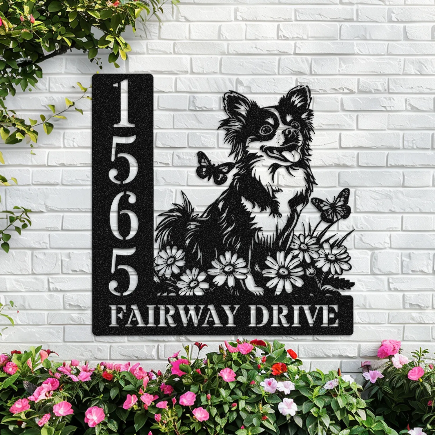 Black metal house sign mounted on a white brick wall showing numbers 1565 and text 'FAIRWAY DRIVE' with a decorative silhouette of a smiling long-haired Chihuahua among daisies and butterflies, pink flowers below.