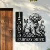 Black metal house sign mounted next to a wooden door showing address numbers 1565 and the street name FAIRWAY DRIVE with a decorative puppy, flowers and butterfly design