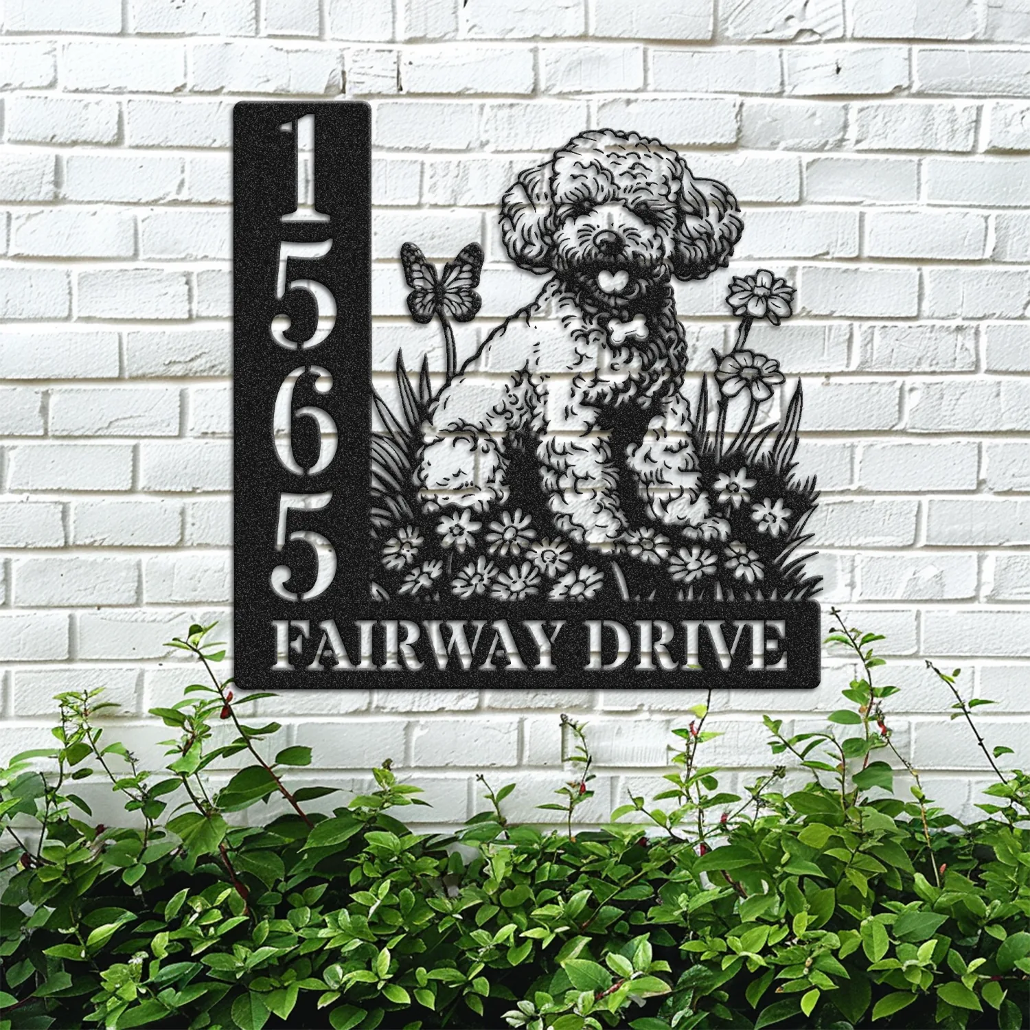 Black metal house sign mounted on a white brick wall showing numbers 1565 and the text FAIRWAY DRIVE with a carved poodle sitting among flowers and a butterfly, green shrubs below.